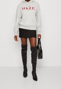 Felpa grigia con logo "VOGUE" in rosso, abbinata a una mini gonna nera e stivali alti neri. Una borsa nera completa l'outfit.