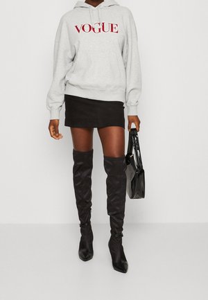 Felpa grigia con logo "VOGUE" in rosso, abbinata a una mini gonna nera e stivali alti neri. Una borsa nera completa l'outfit.