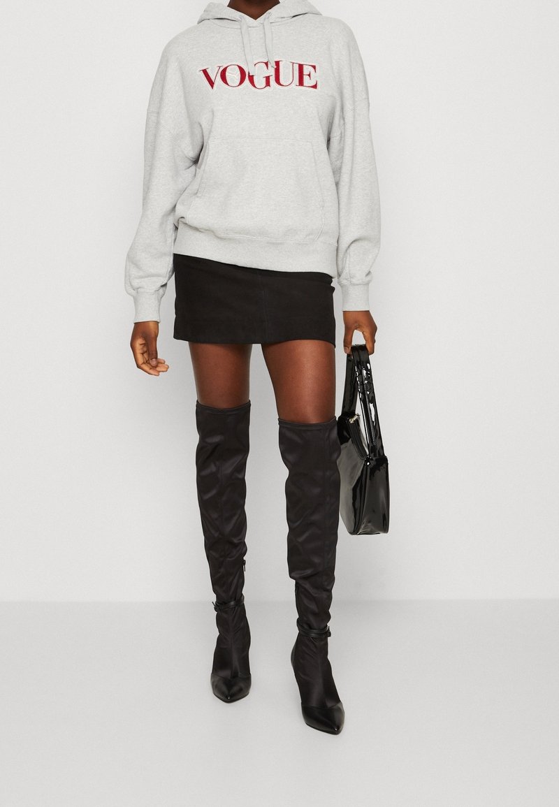Felpa grigia con logo "VOGUE" in rosso, abbinata a una mini gonna nera e stivali alti neri. Una borsa nera completa l'outfit.