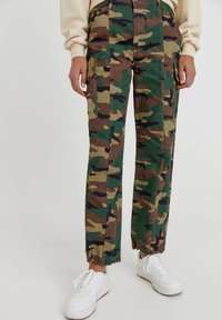Camo cargo pants in groen, bruin en zwart. Bevat zijzakken, een rechte snit en elastische boorden. Gecombineerd met witte sneakers.