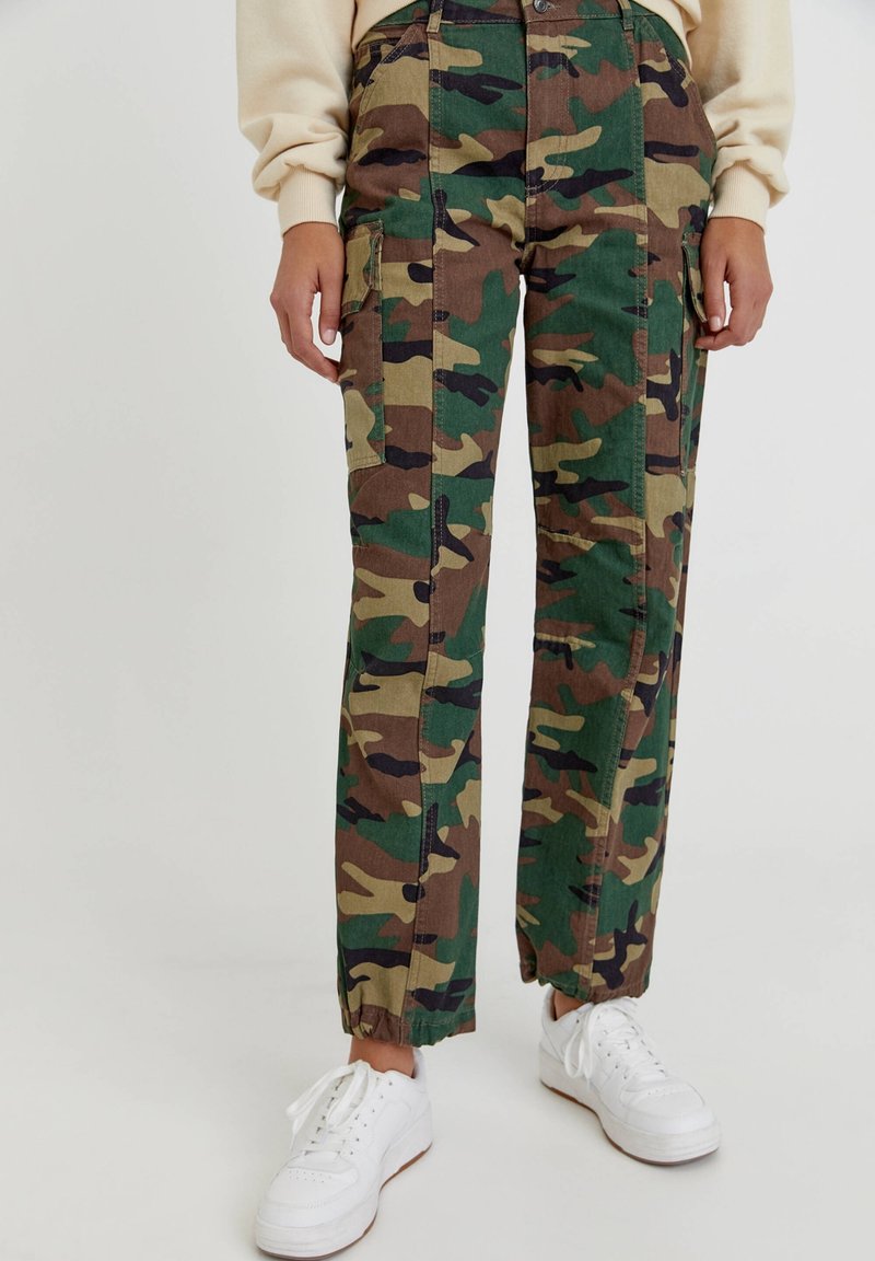 Camo cargo pants in groen, bruin en zwart. Bevat zijzakken, een rechte snit en elastische boorden. Gecombineerd met witte sneakers.