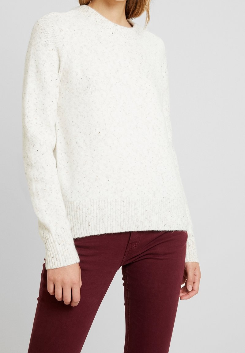 Femme portant un pull en maille texturée blanc et un pantalon slim rouge foncé, debout devant un fond gris clair uni.