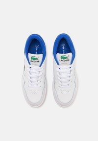 Pár bílých tenisek Lacoste s modrou vnitřní podšívkou, bílými tkaničkami a zeleným krokodýlem na jazyku a na boku.