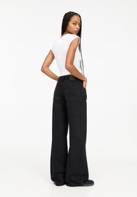 Schwarze weit geschnittene Jeans mit hohen Bund, ausgestattet mit Gesäßtaschen und einem strukturierten Stoff. Kombiniert mit einem taillierten weißen Oberteil.