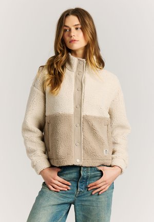 Jonge vrouw die een beige en crème fleecejack met drukknopen draagt en een blauwe spijkerbroek, handen op de heupen, zijwaarts kijkend.