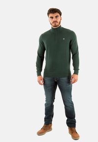 Maglione verde lavorato a maglia con colletto alto e zip a metà, abbinato a jeans in denim blu e stivali beige con lacci. Dettaglio logo discreto sul petto.