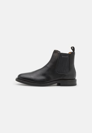Stivale Chelsea in pelle nera con pannello elastico ai lati, linguetta sul tallone e tacco basso, mostrato di profilo su sfondo bianco.