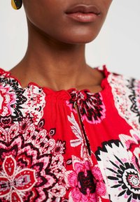 Primer plano de una persona que lleva una blusa roja con un estampado con diseños florales en rosa, blanco y negro, y con un escote en forma de lágrima.