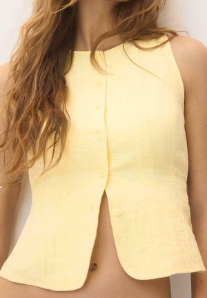 Femme aux longs cheveux ondulés portant un haut sans manches jaune pâle boutonné, montrant le ventre et un bracelet en or à son bras.