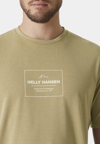 Světle olivově zelené tričko z hladkého materiálu. Obsahuje obdélníkovou bílou nálepku s textem, umístěnou uprostřed hrudi. Krátké rukávy a kulatý výstřih.