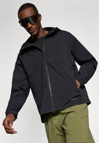Veste imperméable noire avec fermeture éclair, poignets élastiques et capuche, portée par-dessus une chemise noire. Le modèle porte de grandes lunettes de soleil sombres et un short vert.