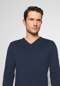 Pulover bleu marin cu decolteu în V, cu mâneci lungi, textură cu nervuri și croială ajustată, fabricat dintr-un material moale. Stil minimalist fără detalii decorative.