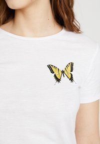 Camiseta de algodón blanca con una mariposa bordada en negro y amarillo en el lado izquierdo del pecho. Cuello redondo y mangas cortas.