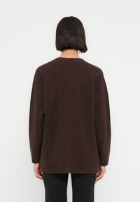 Καφέ πλεκτό πουλόβερ με φαρδιά εφαρμογή, υφή με ρίγες, στρογγυλή λαιμόκοψη και μακριά μανίκια, φορεμένο πάνω από σκούρο παντελόνι, με θ έα από πίσω.