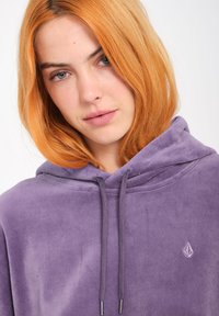 Felpa con cappuccio viola in morbido e peloso materiale, dotata di tasca frontale e lacci; presenta un piccolo logo sul petto.