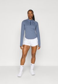 Lett blå langermet idrettstopp med glidelås i kragen, kombinert med hvite shorts. Modellen har på seg hvite sokker og sko.