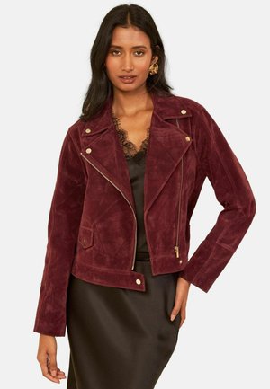 Femme portant une veste biker en daim bordeaux avec des détails dorés sur une robe en satin noire bordée de dentelle, debout devant un fond uni.