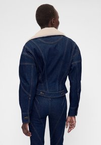 Tumesinine teksajakk pehme beežika kraega. Detailideks on õmblused ning nupud vöökohal ja mansettidel. Vaade tagant.