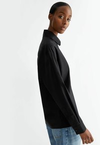 Camisa negra de manga larga con cuello clásico, textura suave y diseño entallado. Presenta detalles de botones en los puños y una abertura lateral.