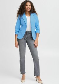 Světlomodrý blazer s ohrnutými rukávy, bílý top a šedé džíny. Modelka stojí v béžových lodičkách před jednobarevným pozadím.