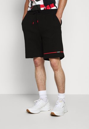 Mann trägt schwarze Shorts mit rotem Rand und weiße Sneakers, steht mit den Händen in den Taschen vor weißem Hintergrund.