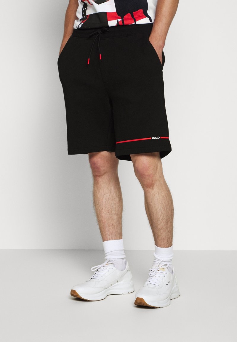 Homme portant un short noir avec une bordure rouge et des baskets blanches, debout les mains dans les poches sur fond blanc.