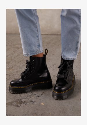 Botas de cuero negro hasta el tobillo con suela gruesa, puntera redonda, cintas de raso, costuras amarillas y una tira en la parte trasera.