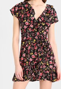 Vestido negro con flores, con mangas rizadas, que presenta flores rosas y amarillas. El diseño incluye un escote en V y una cintura ceñida con un lazo.