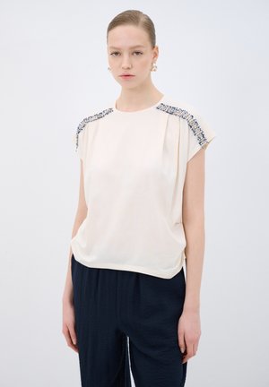Haut à manches courtes couleur crème avec des perles texturées bleu marine le long des épaules, offrant une coupe décontractée et un tissu doux.