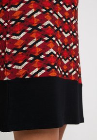 Robe rouge à motifs présentant un design géométrique en orange et blanc avec un ourlet noir. Fabriquée en tissu avec une texture visible.