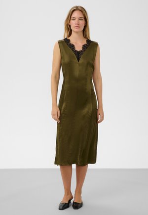 Femme aux longs cheveux blonds portant une robe midi sans manches vert olive avec un col en dentelle noire, debout devant un fond gris clair uni.