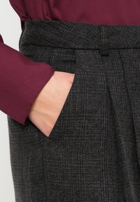 Hand in der Tasche von hochtaillierten, dunkelgrauen karierter Hose, kombiniert mit einem bordeauxfarbenen Langarmoberteil, das die Stoffstruktur und den Schnitt zeigt.