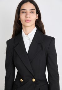 Čierny dvojradový blazer z pevného materiálu, srdce zlato gombíkov, ostré klopy a biela košeľa pod ním, hladká textúra.