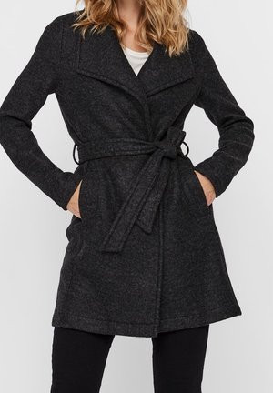 Femme portant un manteau en laine gris foncé ceinturé avec de larges revers, mains dans les poches, associé à un pantalon noir et un haut blanc en dessous.