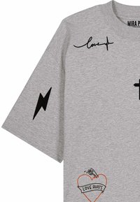 T-shirt gris clair avec un éclair noir sur la manche, « love » avec un tracé de rythme cardiaque près du col, et un cœur avec un pistolet et le texte « LOVE HURTS » devant.