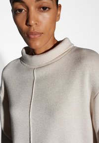 Maglione a maglia beige con un col alto e ripiegato, caratterizzato da una sottile lucentezza e una texture liscia. Include un dettaglio di cucitura verticale.