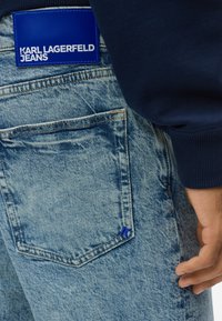 Šviesiai mėlynos džinsinės kelnės su tamsiai mėlynu juosmeniu. Pridėta mėlyna etiketė su užrašu "KARL LAGERFELD JEANS." Kelnės turi nugarinį kišenę su mėlynu akcentu.