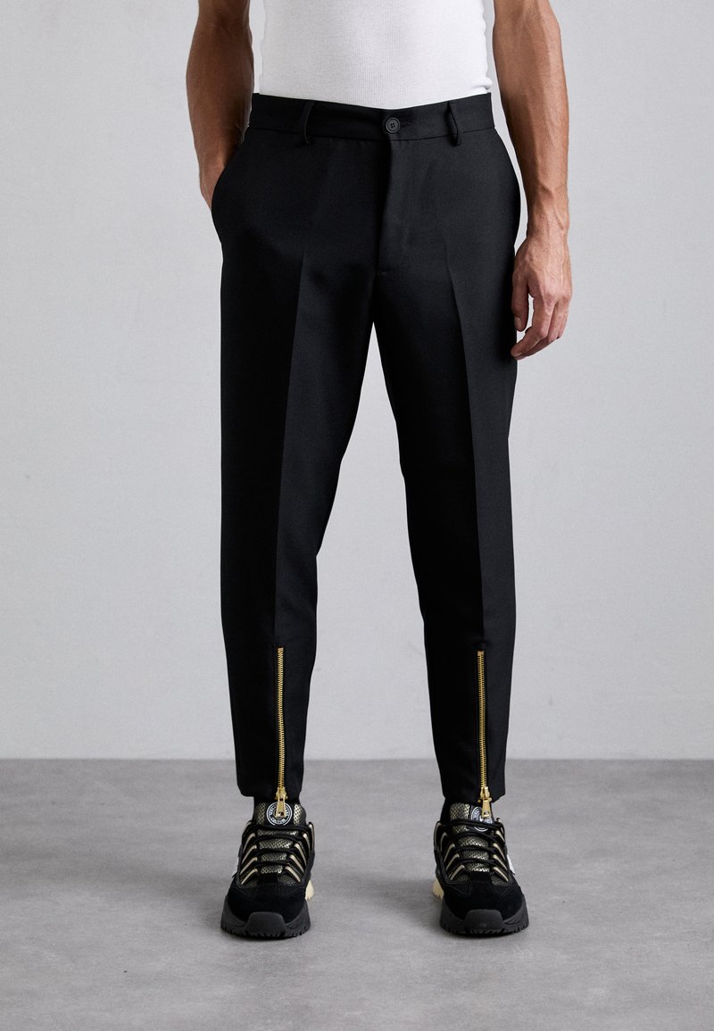 Zwarte op maat gemaakte broek met een gladde stof, voorzien van ritsen aan de voorkant bij de enkels en een slim fit ontwerp. Gepaard met zwarte en gouden sneakers.