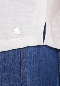 Weißes Leinenhemd mit einem runden silbernen Logo-Knopf am Saum und seitlichen Schlitzen, kombiniert mit blauer Jeansware, die Nähdetails zeigt.