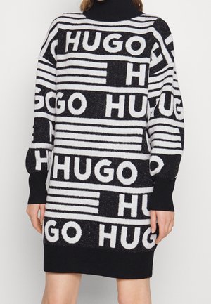 Black and white turtleneck sweater dress featuring repeating "HUGO" text and horizontal stripes, worn by a person with hands relaxed at sides.