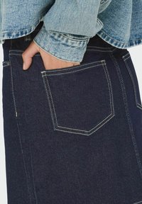 Ciemnoniebieska spódnica dżinsowa z tylną kieszenią, z kontrastowym przeszyciem i prostym krojem. Na wierzchu znajduje się jasnoniebieska kurtka dżinsowa.