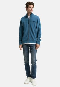 Trekker fleece jack in teal, met een hoge kraag, een borstzak en geribde manchetten. Gecombineerd met blauwe jeans en witte sneakers.