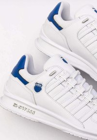 Białe sportowe sneakersy z niebieskimi akcentami, siateczkowym językiem i wieloma pętlami na sznurowadła. Zawierają srebrne logo "K-Swiss" z boku.