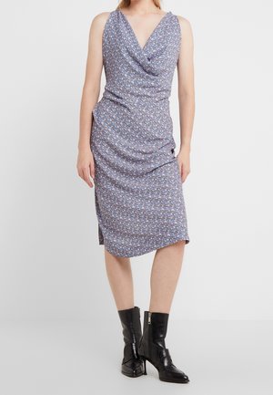 Kobieta ubrana w bezrękawnikową, sięgającą kolan fioletową sukienkę w kwiaty z dekoltem z drapowaniem oraz czarne skórzane botki za kostkę z zamkami po bokach.