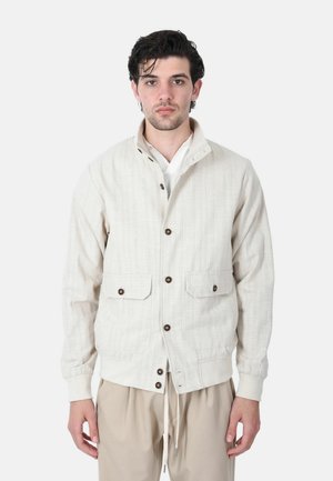Uomo che indossa una giacca beige chiaro con bottoni e tasche a patta su una camicia bianca e pantaloni beige, in piedi davanti a uno sfondo semplice.