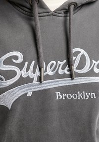 Hanorac gri cu un logo brodat "SuperDry" și textul "Brooklyn". Dispune de sireturi și un material moale, texturat.