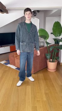 Jaqueta de ganga azul escuro com dois bolsos frontais, combinada com jeans azuis e ténis brancos. Ambiente interior com grandes plantas verdes.