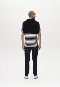 T-shirt a righe con maniche corte indossato sotto un pullover blu scuro. La modella indossa jeans blu scuro e scarpe di colore chiaro. Sfondo bianco semplice.