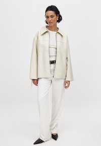 Kremo spalvos dirbtinė odinė striukė su apykakle, užtrauktuku priekyje ir šoninėmis kišenėmis, dėvima ant baltos bluzės ir kreminės spalvos kelnėmis, baigta juodais kulnais.