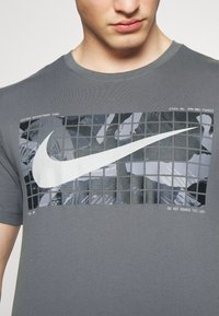 Grå t-shirt med en stor vit Nike-swoosh på en svart och grå rutnätsgrafik med ett kamouflagemönster. Rund hals, kort ärm.